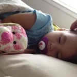 Toddler asleep with a pacifier and stuffed toys, a parent's hand near their head: sick day activities for toddlers