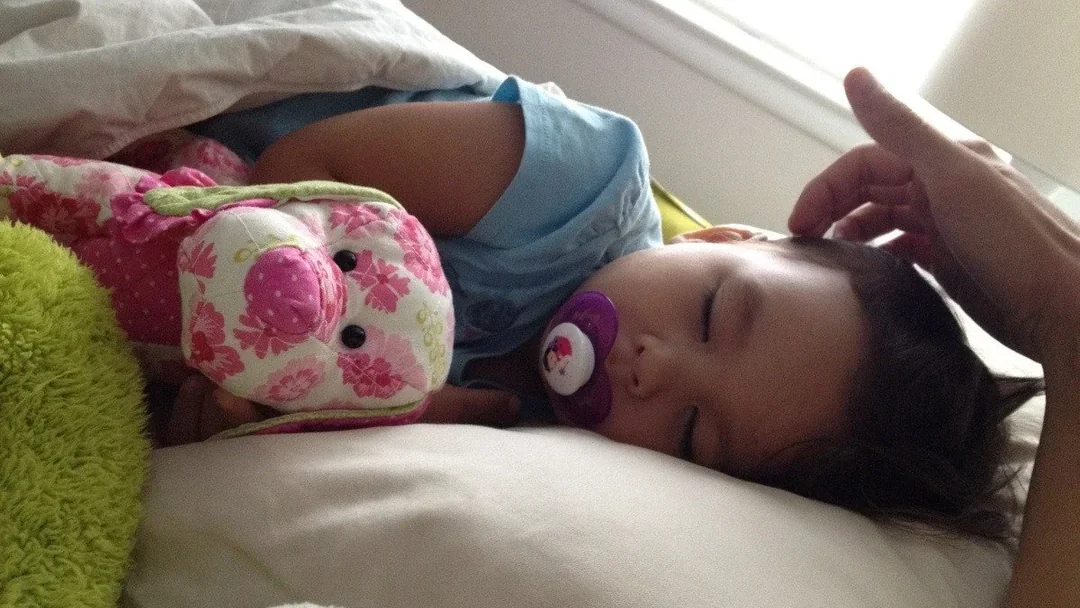 Toddler asleep with a pacifier and stuffed toys, a parent's hand near their head: sick day activities for toddlers