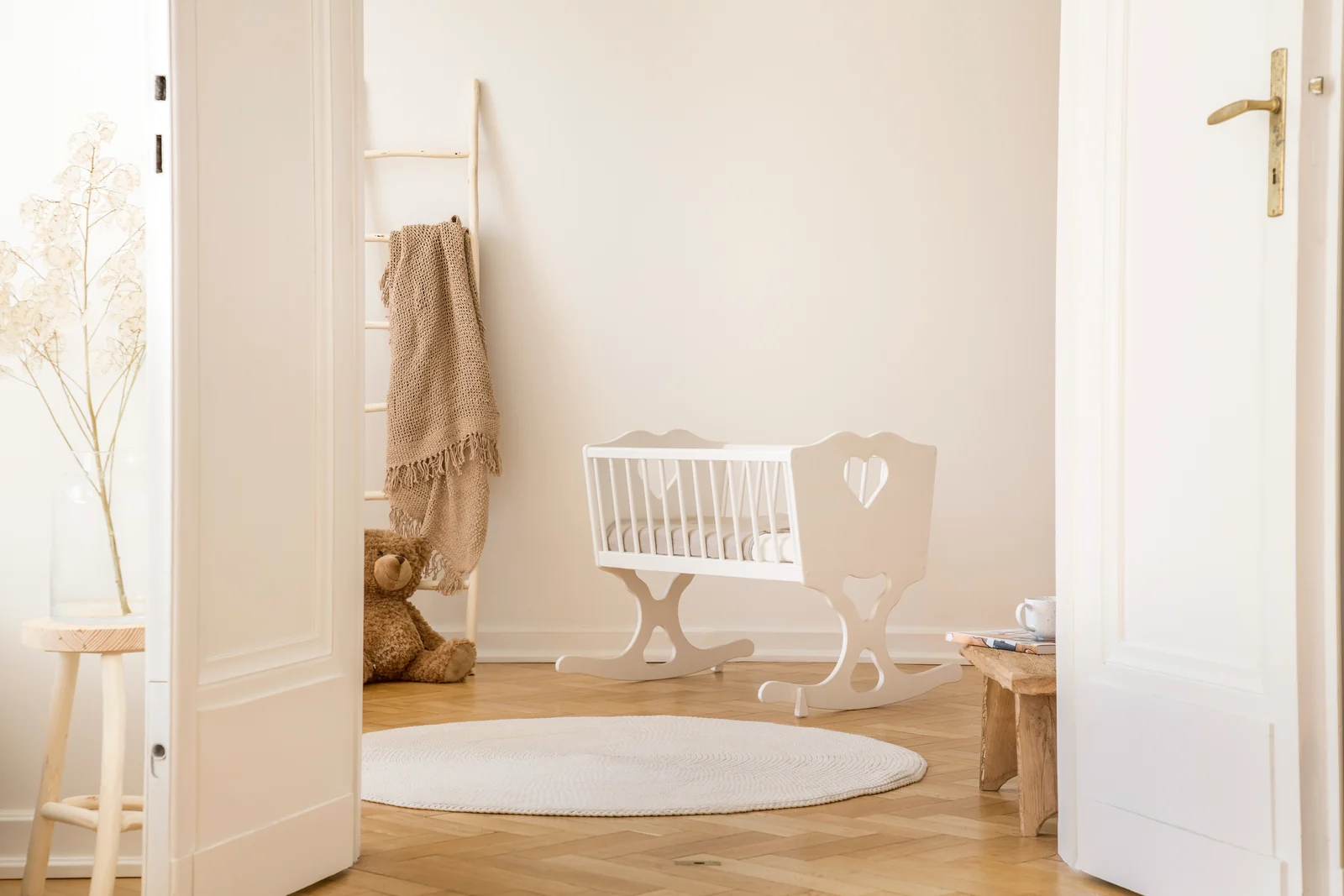 A small white cradle with heart cutouts on a round rug in a plain, bright nursery, seen through an open doorway