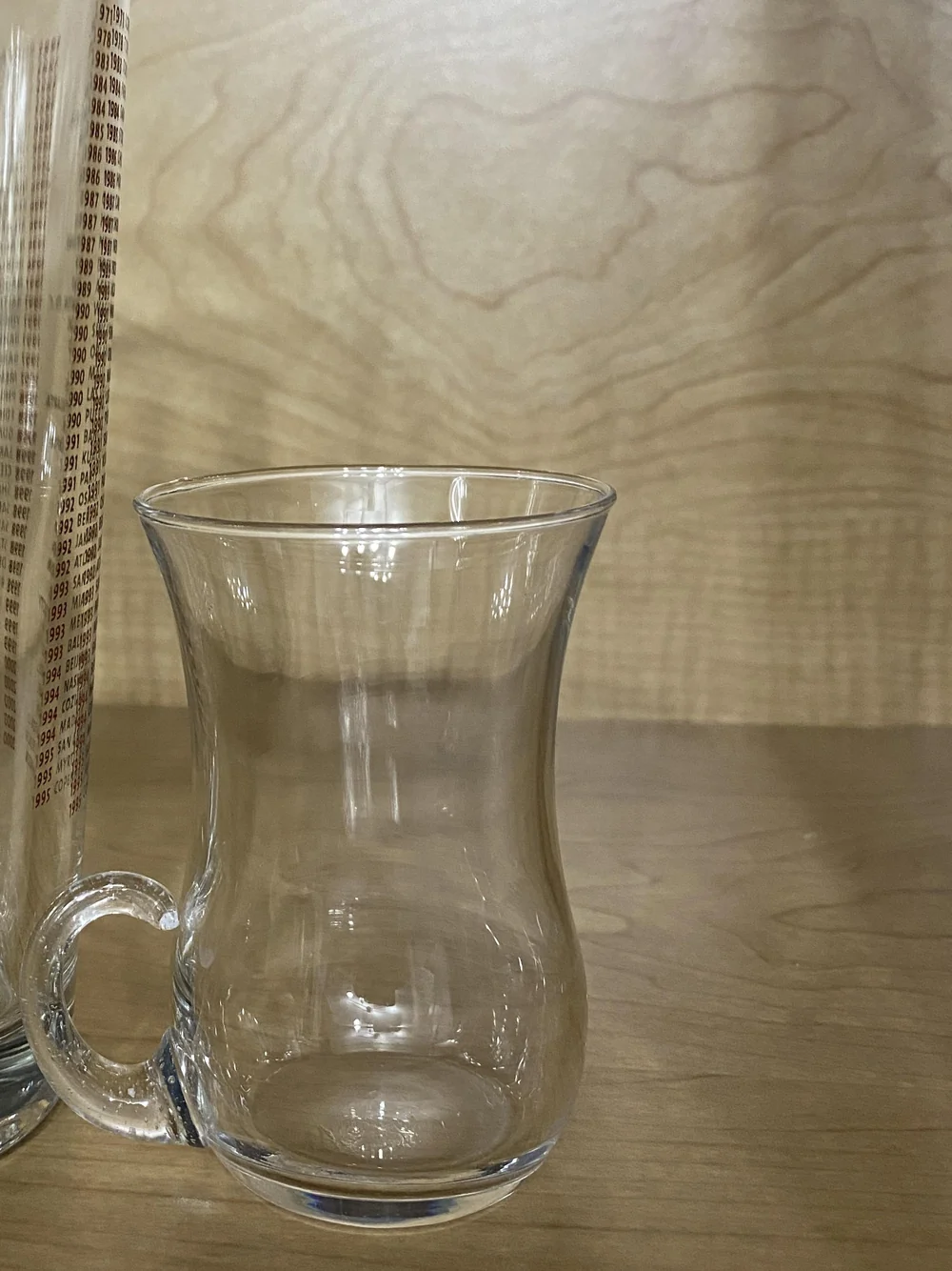 A small clear glass cup with a curved handle on a light wood shelf