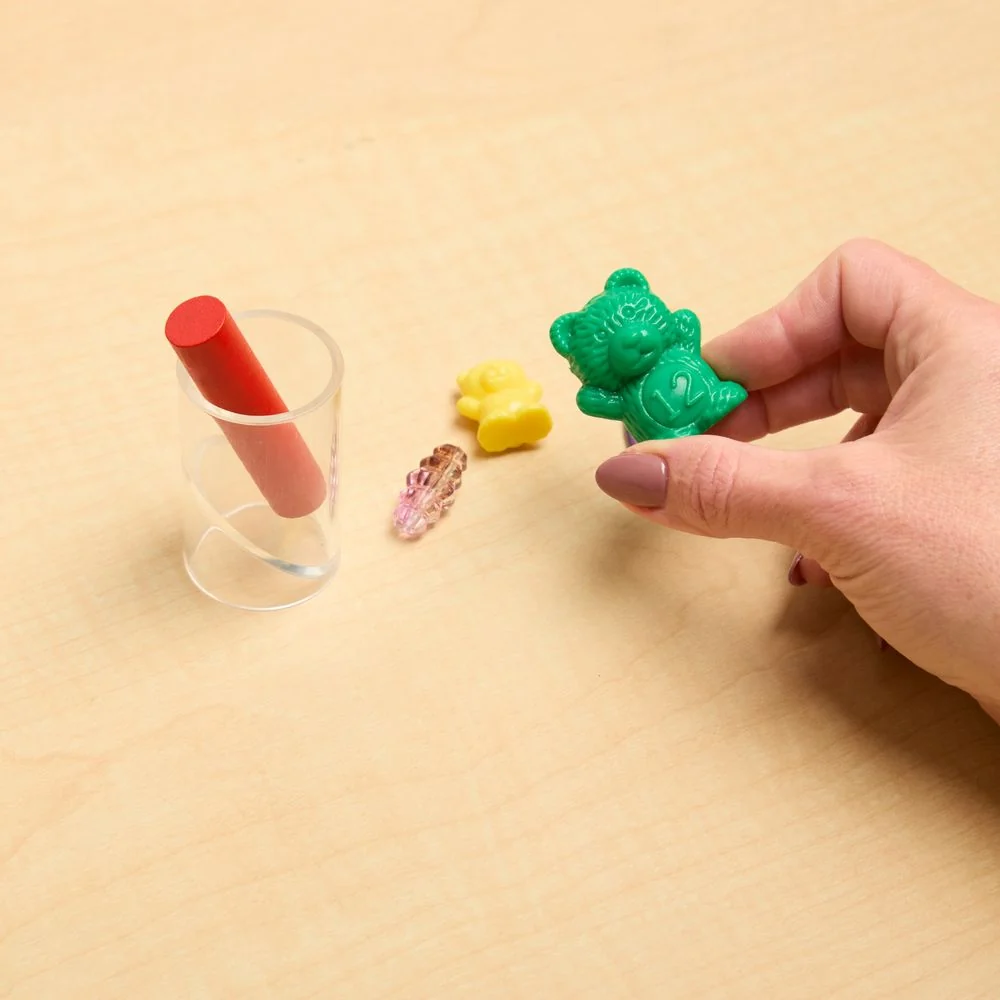 A clear small-parts test cylinder on a wooden table with a red peg inside, two small plastic bears and a bead, one bear held in a hand