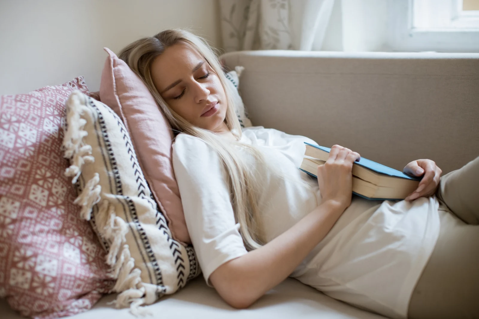 A tired woman dozes on a sofa with an open book resting in her hands