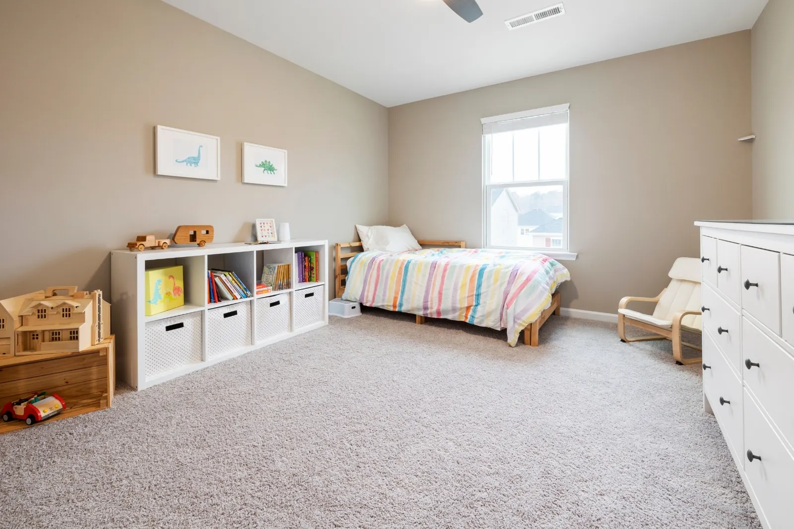 A tidy children's bedroom with a low wooden bed, a white cube shelf of books and wooden toys, and a small armchair