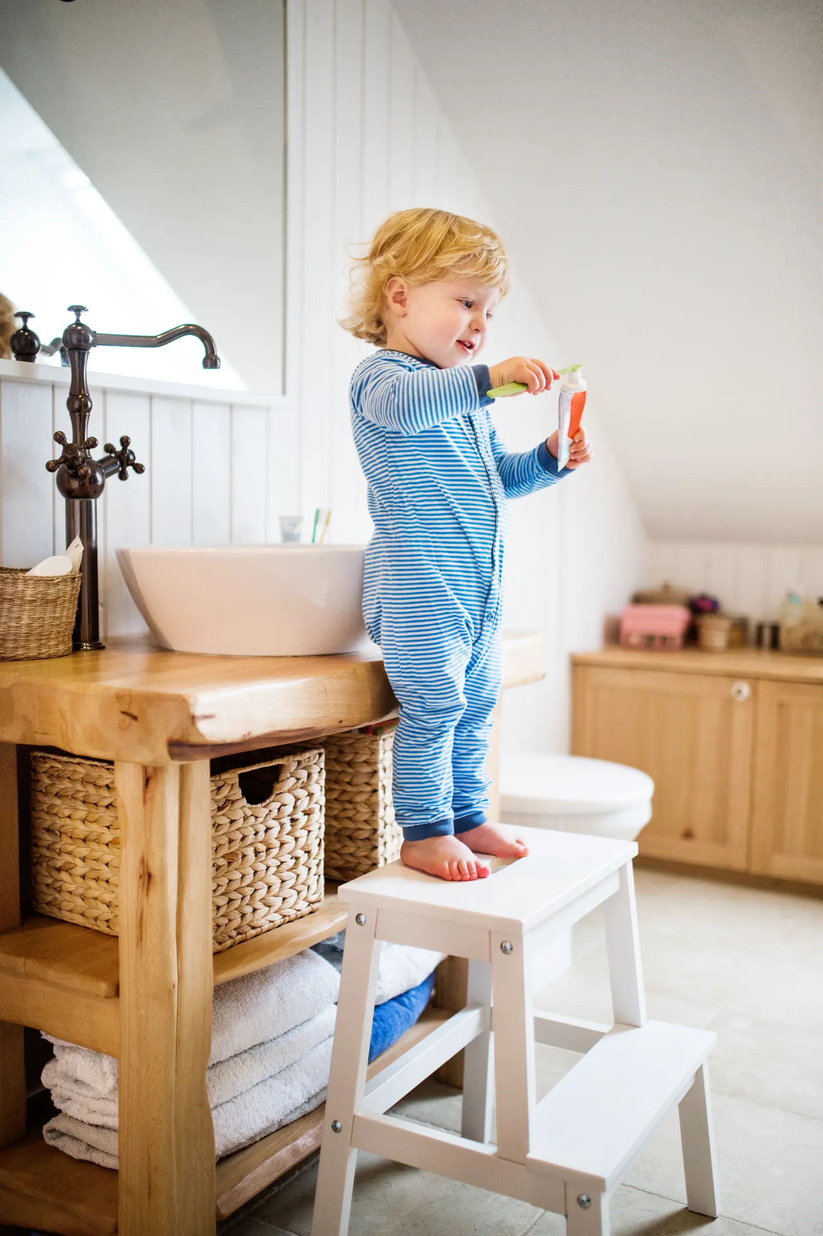 A toddler on a step stool washes their hands at a sink while a parent stands beside them