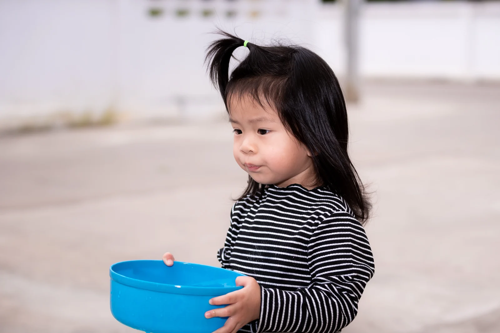 Toddler girl in a striped top carrying a blue plastic bowl in both hands, looking off to one side