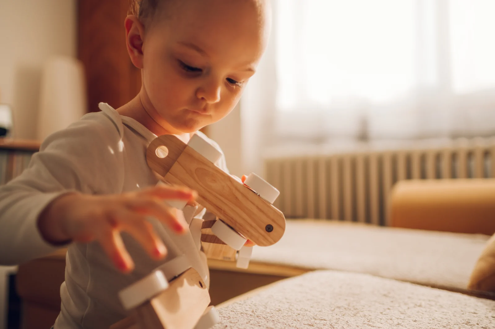 Toddler holding a chunky wooden toy vehicle with both hands, looking down at it by a sunny window