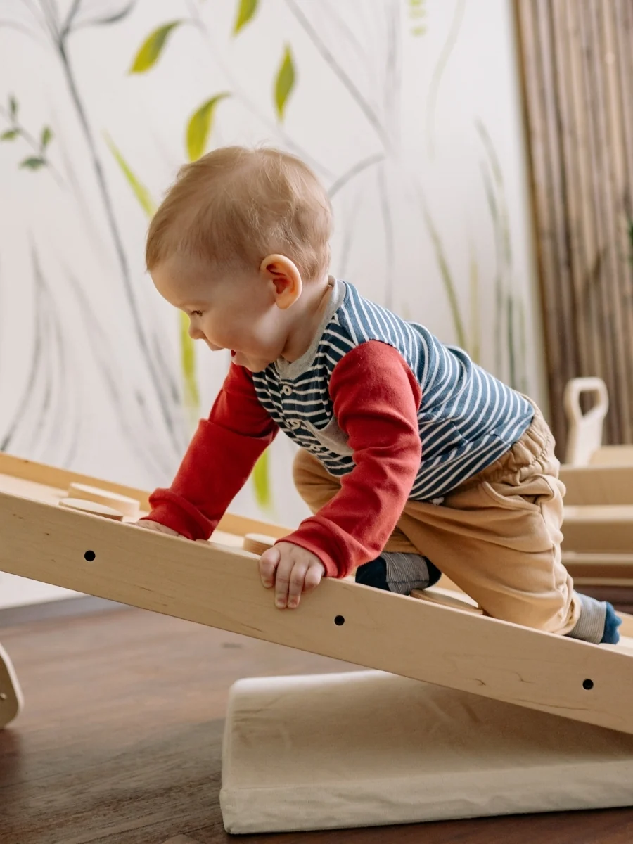 A toddler climbs the ramp of a low wooden climbing frame indoors