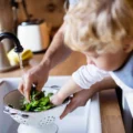 Toddler rinsing spinach in a colander at the kitchen sink while a parent's hand helps, a first step in toddler cooking