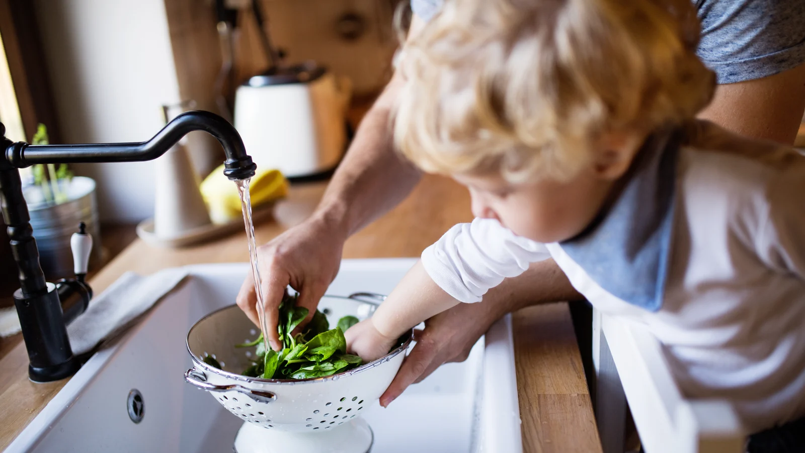 Toddler rinsing spinach in a colander at the kitchen sink while a parent's hand helps, a first step in toddler cooking