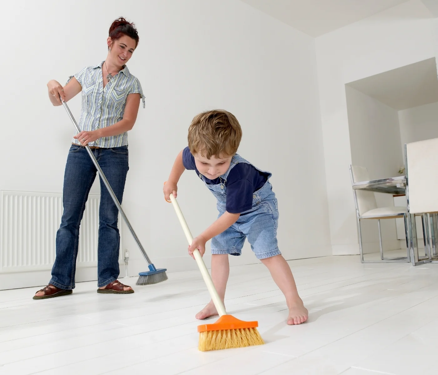 Young child sweeping the floor right beside a parent, each with their own broom