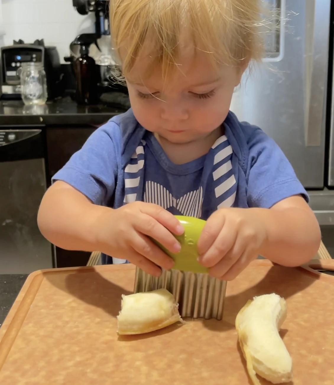 Toddler in a blue shirt pressing a wavy crinkle cutter down on a peeled banana on a cutting board