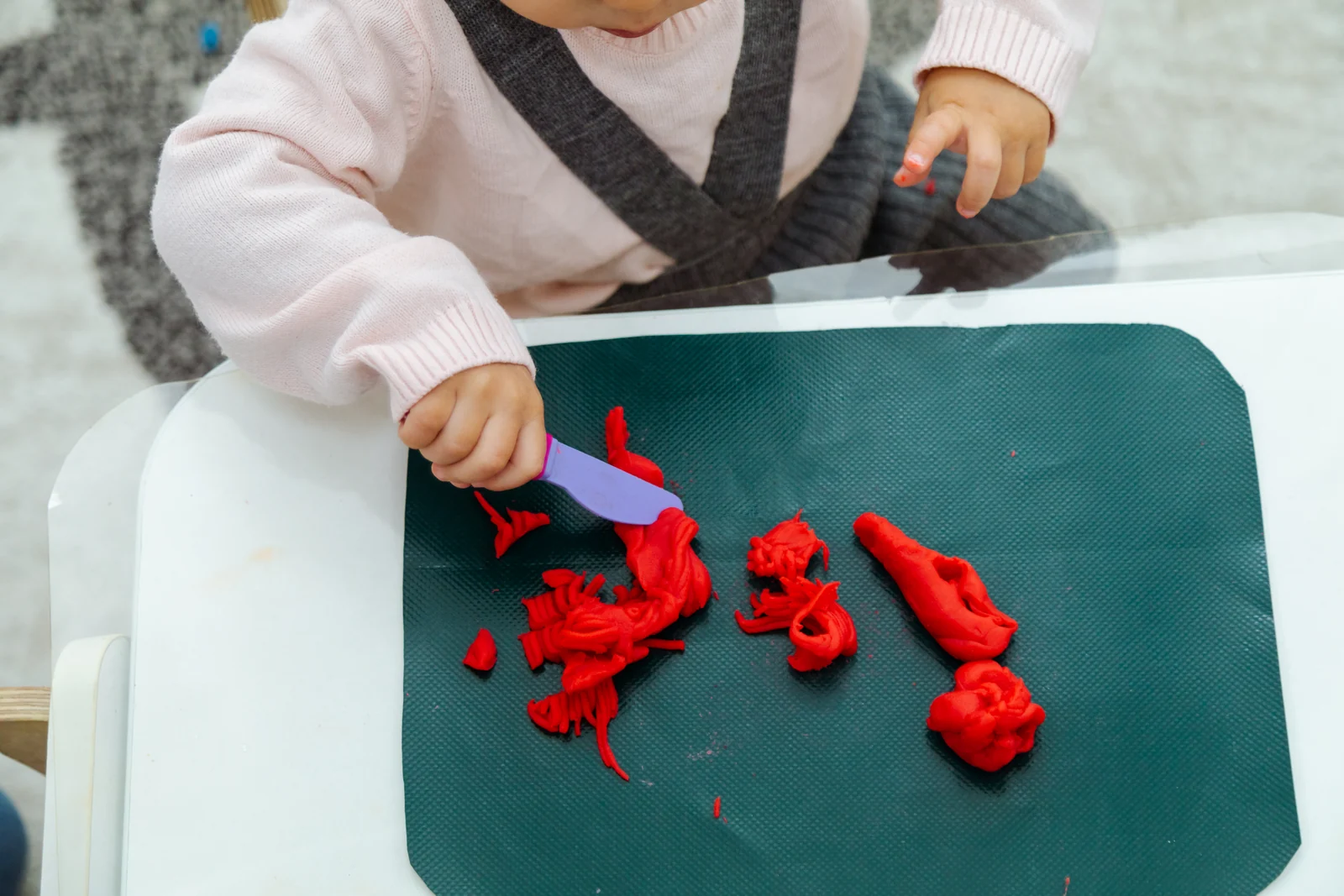 Toddler's hands cutting red playdough with a purple play knife on a dark green mat at a small table