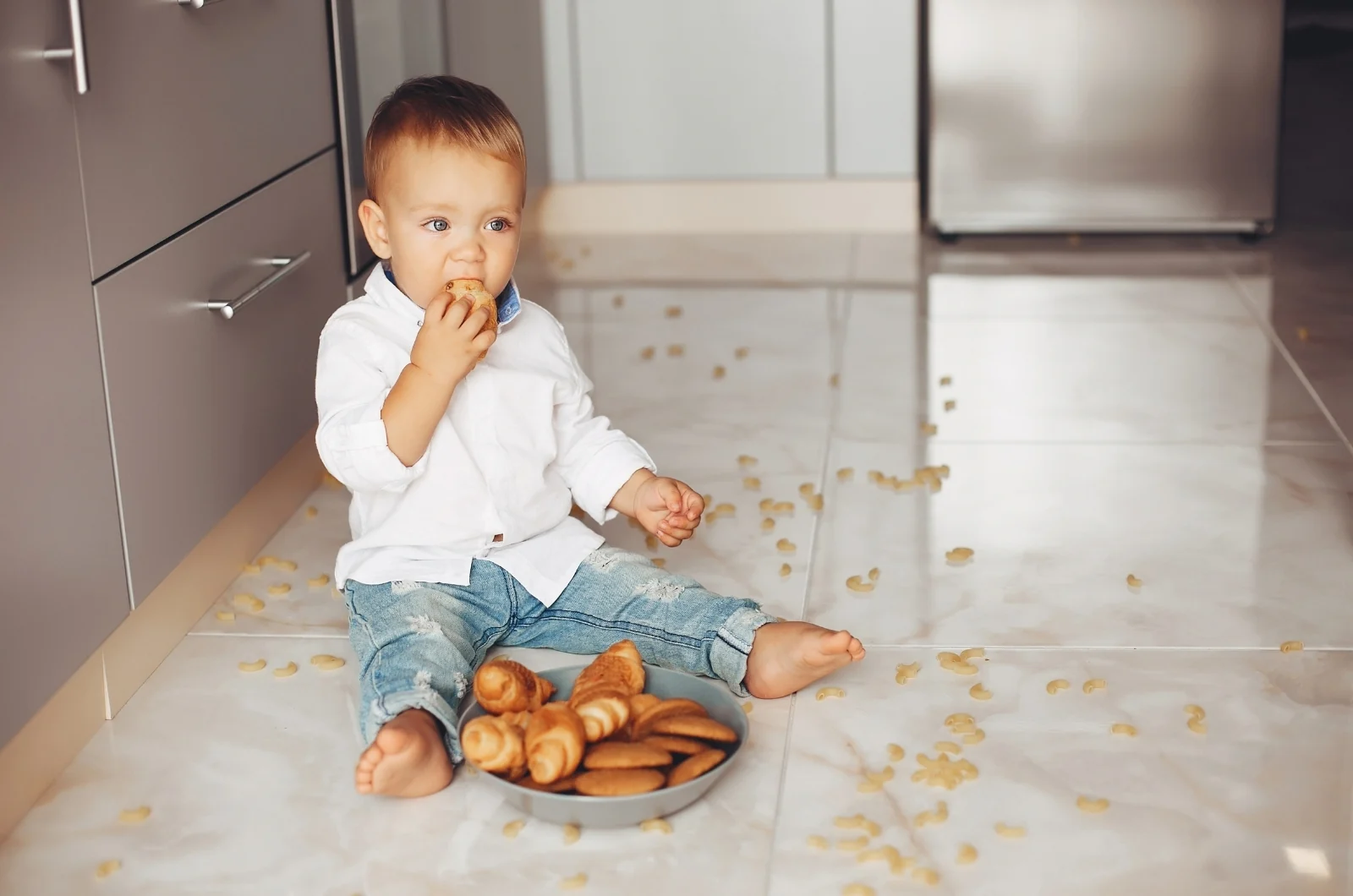 Toddler sitting on a kitchen floor with a bowl of bread rolls, eating a snack among scattered crumbs