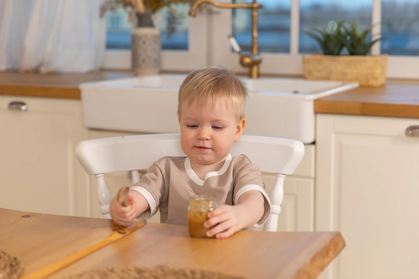 Toddler at a wooden kitchen table, feeding himself from a small jar with a wooden spoon