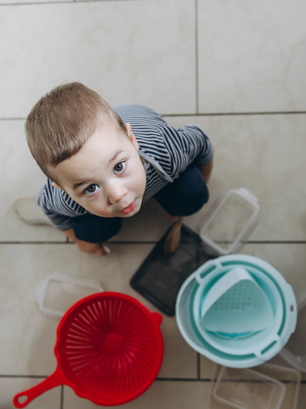 Toddler looking up from a tile floor, surrounded by plastic food containers, a red colander and nesting bowls