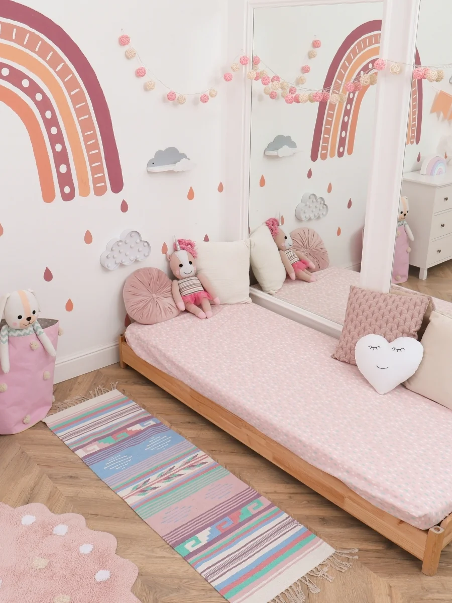 A low floor bed with pink bedding and pillows in a bedroom with a rainbow wall decal and a rug