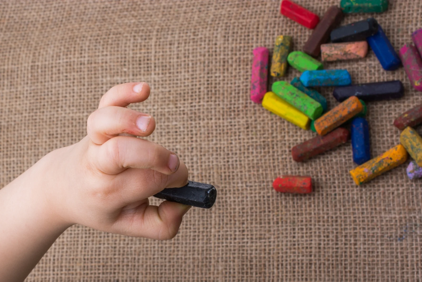 Small child's hand gripping a short, thick crayon, with more crayons scattered on burlap