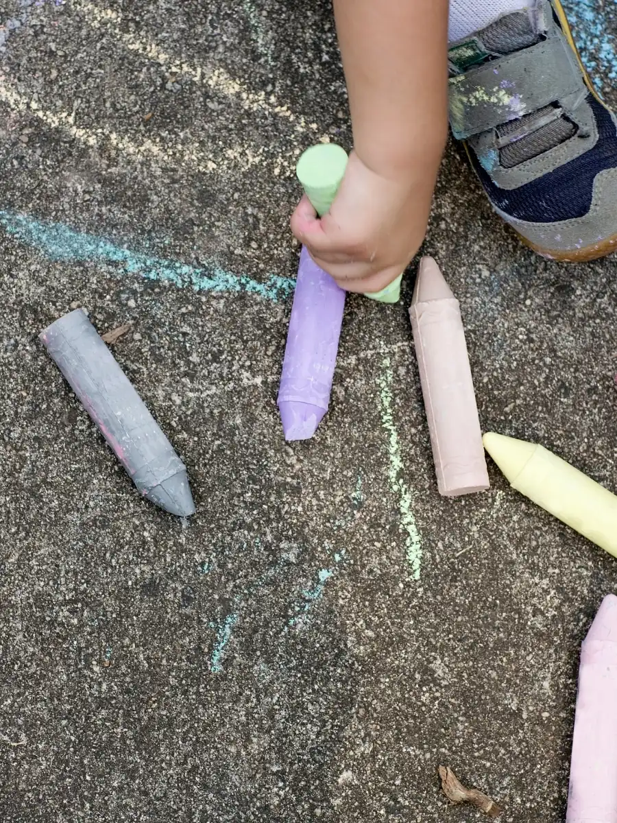 A child's hand holding a thick piece of sidewalk chalk among colorful chunky chalks on pavement