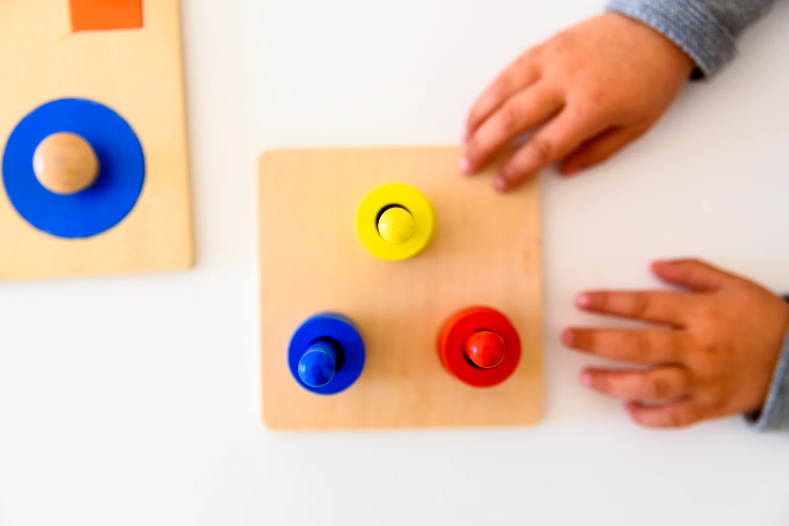 Toddler's hands resting beside a wooden board with three colorful knobbed pieces, seen from above
