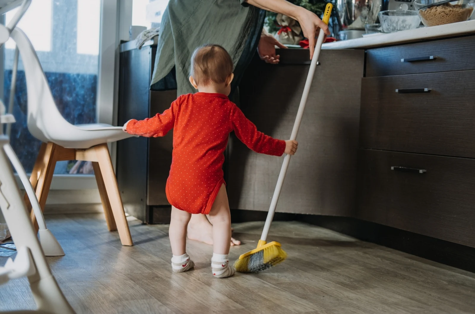 Toddler in a red romper helping an adult sweep the kitchen floor