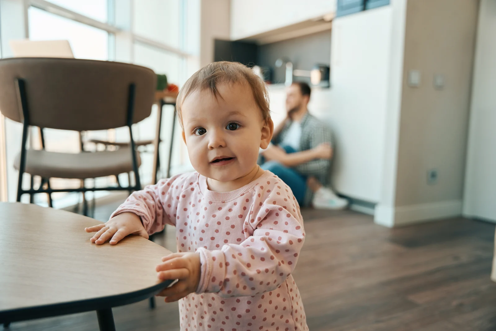 A toddler in a pink polka-dot top standing and holding the edge of a round wooden table in a bright kitchen