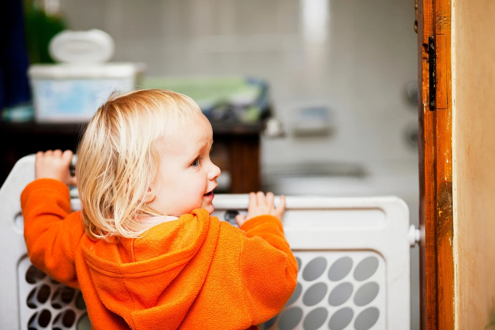 A toddler in an orange hoodie holds the top of a white safety gate and looks into the room beyond