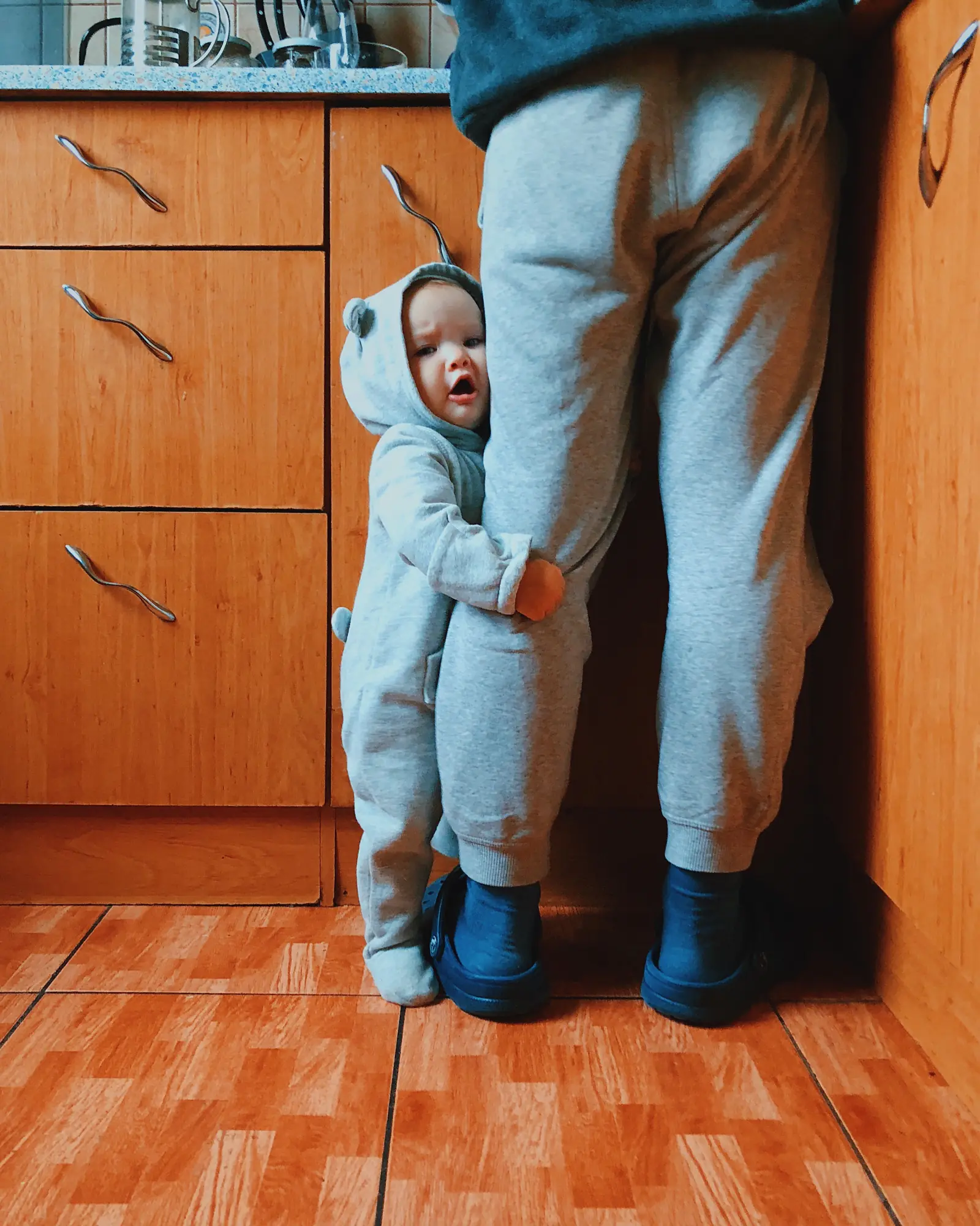 A toddler in a hooded onesie hugging a parent's leg beside the kitchen cabinets
