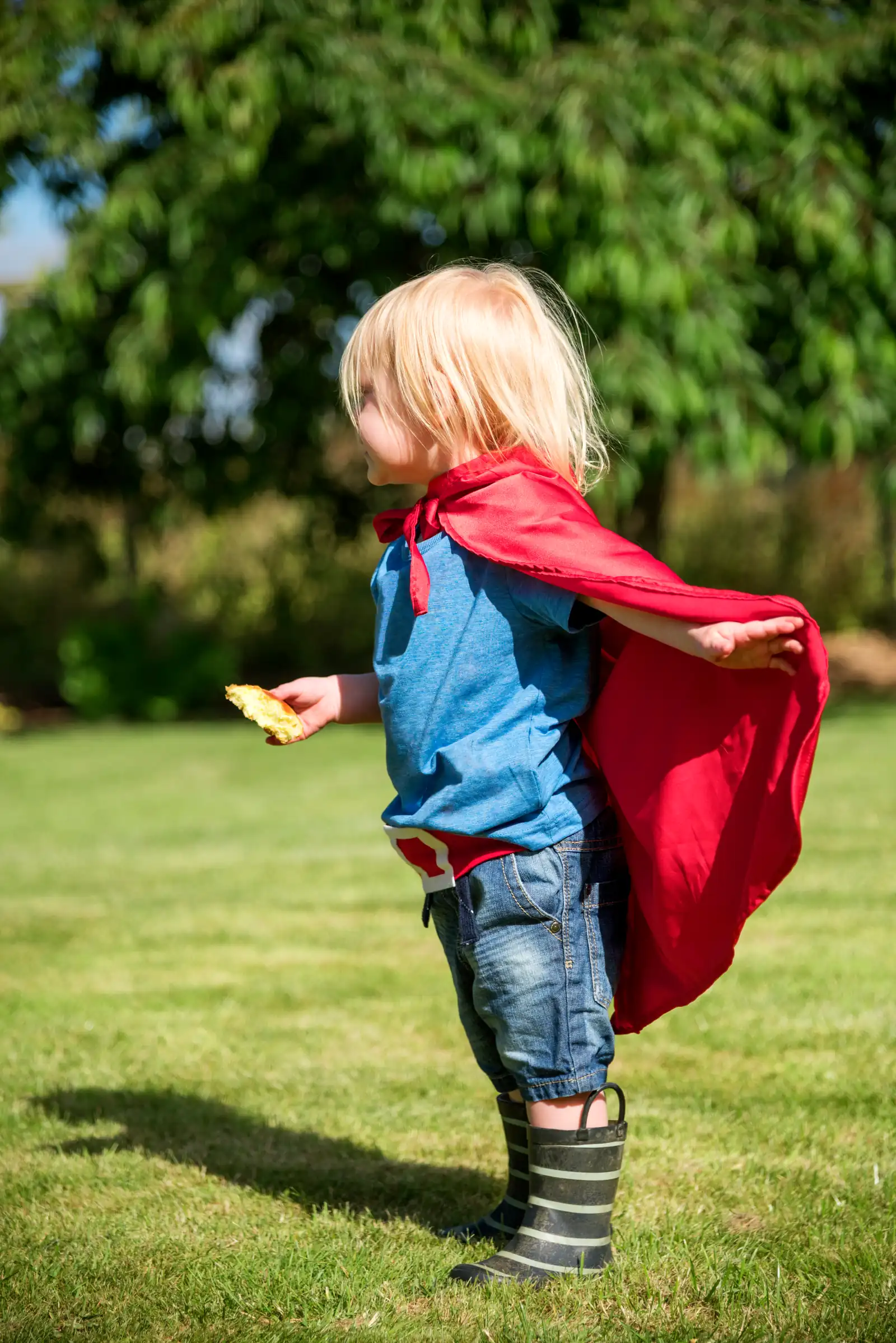 Toddler wearing a plain red cape, arms stretched out, playing pretend in a living room