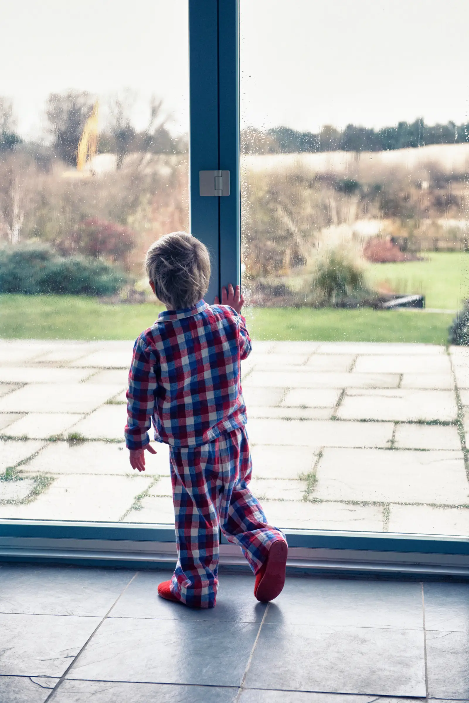A toddler in plaid pajamas standing at a floor-to-ceiling glass door, looking out at a stone patio and garden