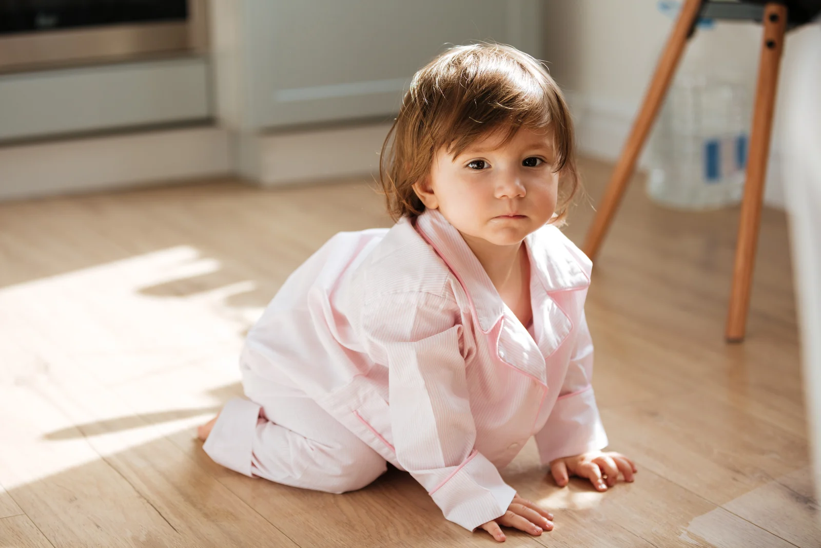 Serious toddler in light pink pajamas kneeling on a sunlit wooden floor and looking at the camera