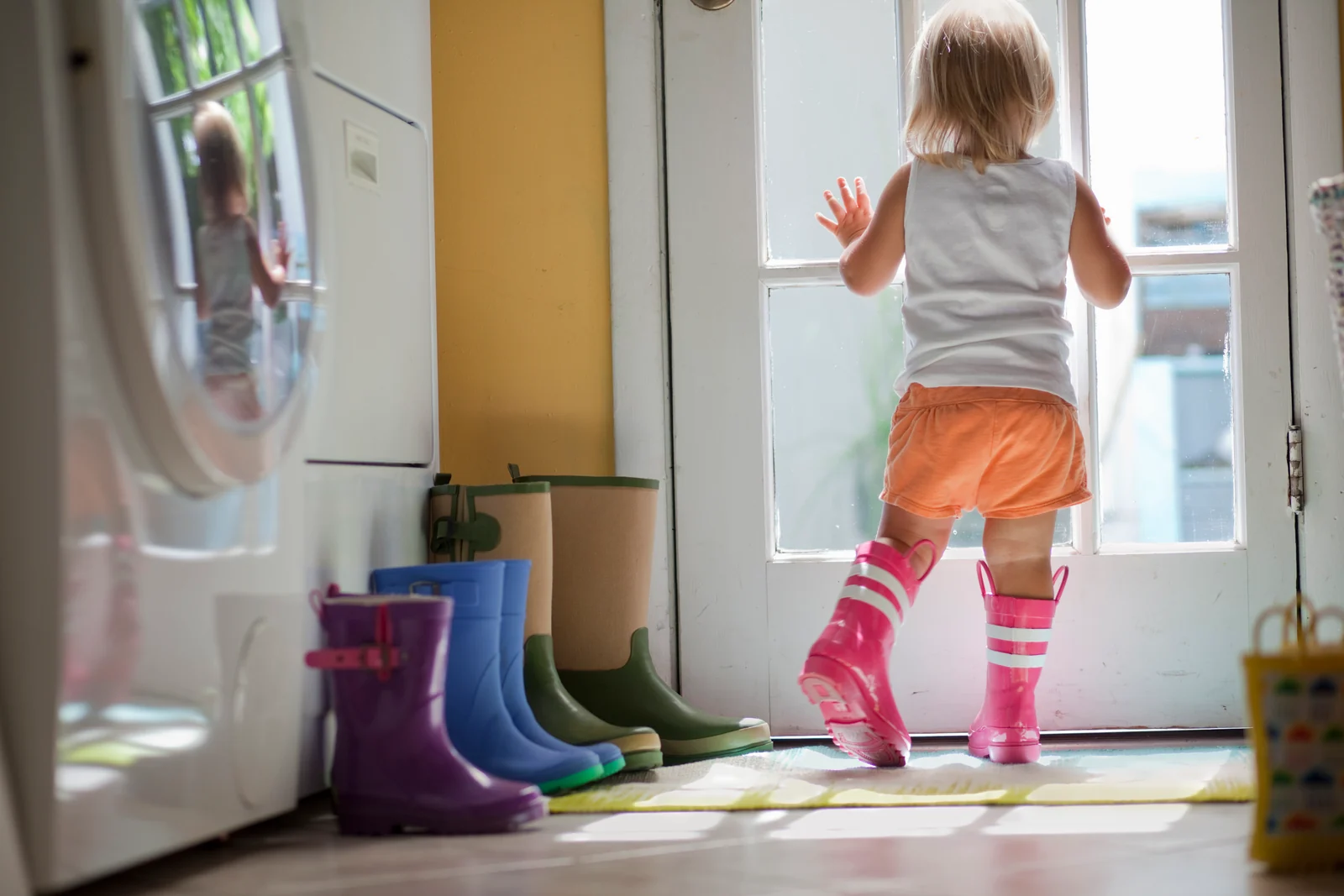 Toddler in pink rain boots at a glass back door, with a row of rain boots lined up by the washing machine
