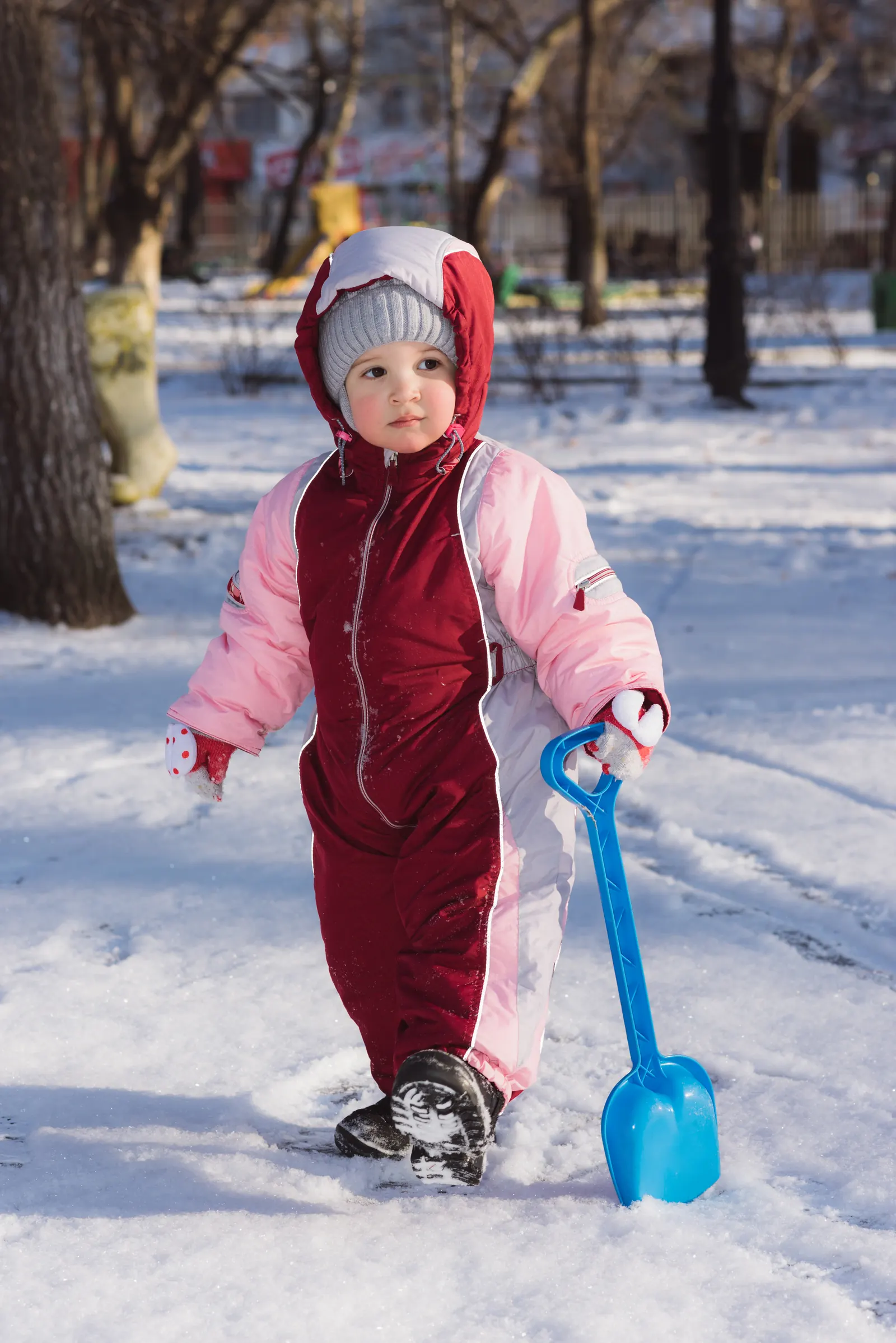 Toddler in a pink and maroon snowsuit holding a blue shovel in a snowy park