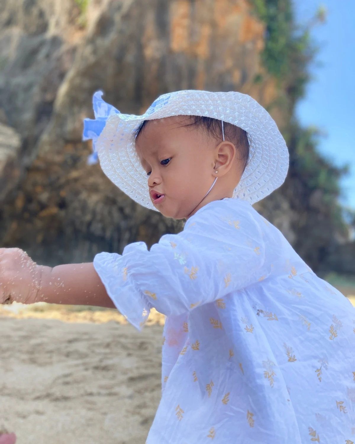 Toddler in a white wide-brimmed sun hat and loose white dress reaching into the sand on a beach