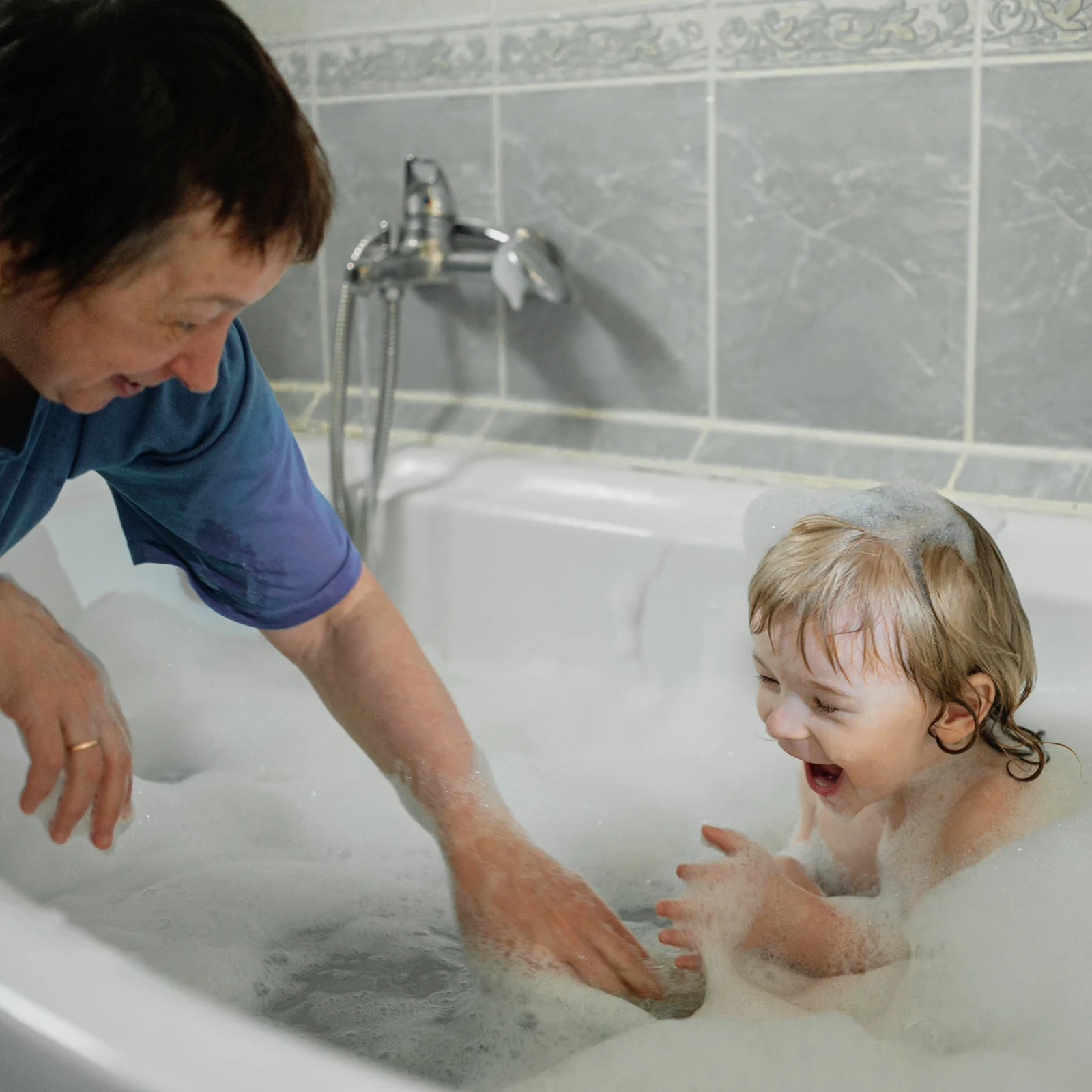 A toddler laughing in a bubble bath while an adult leans in close with a hand in the water beside the child