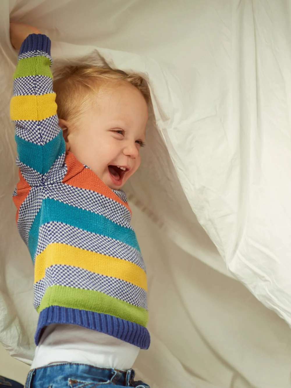 A laughing toddler in a striped sweater plays under a big white sheet, one arm raised