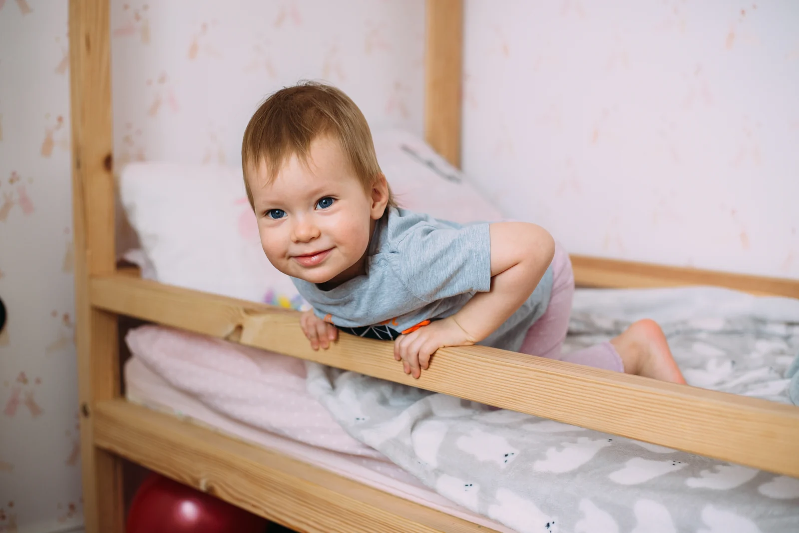 A smiling toddler leaning over the wooden side rail of a low bed with a pillow and a soft blanket
