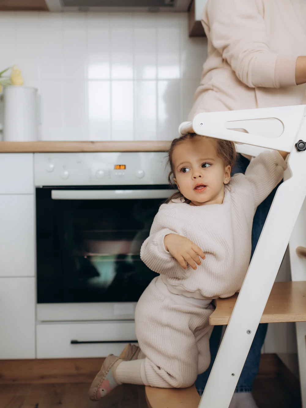 A toddler climbs a white learning tower at the kitchen counter while an adult stands behind