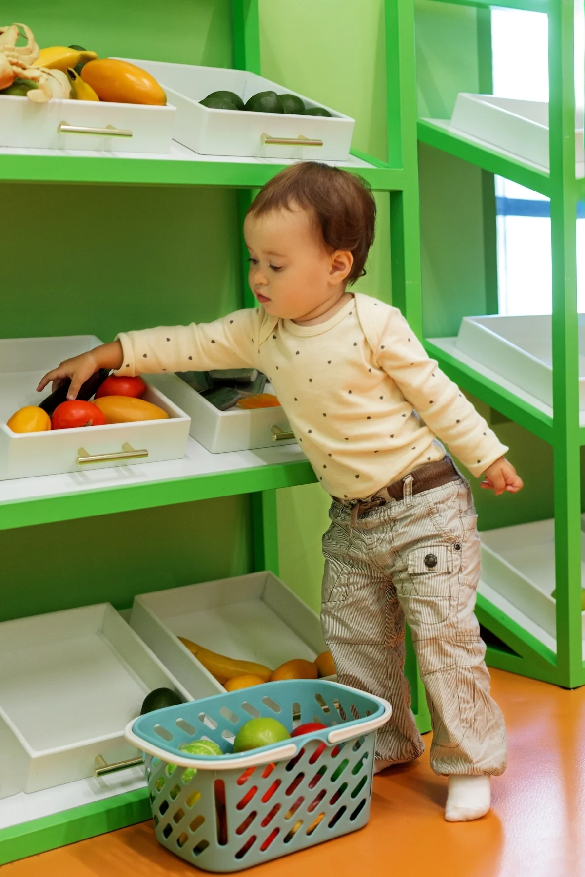 A toddler lifts a small basket off a low open toy shelf