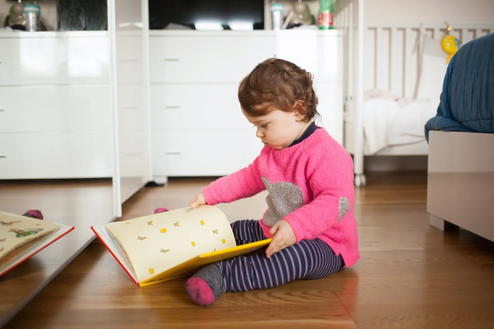 A toddler sits on a rug turning the pages of a sturdy board book