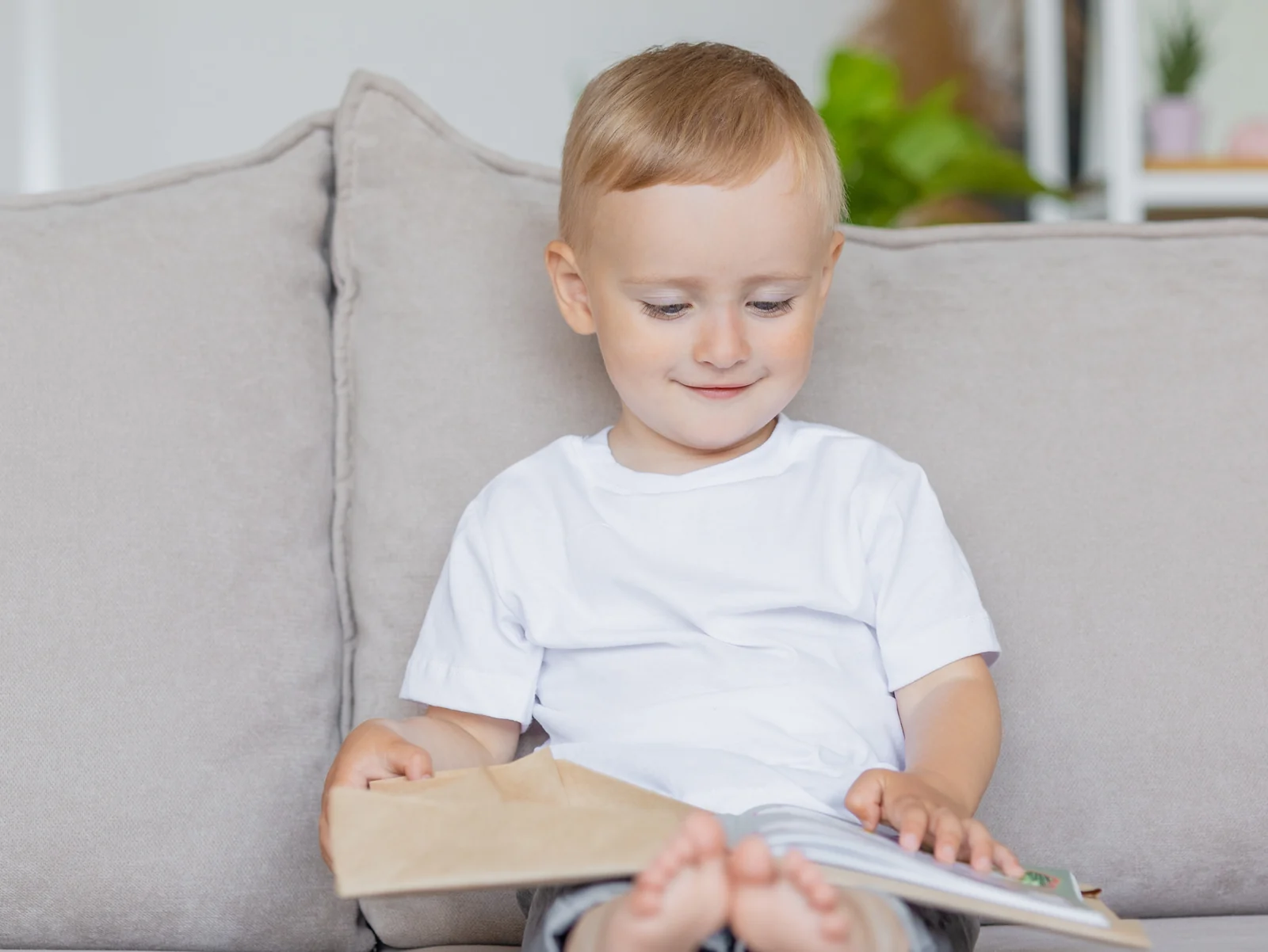 Toddler sitting on a gray couch with a large open book on his lap, looking down at the pages