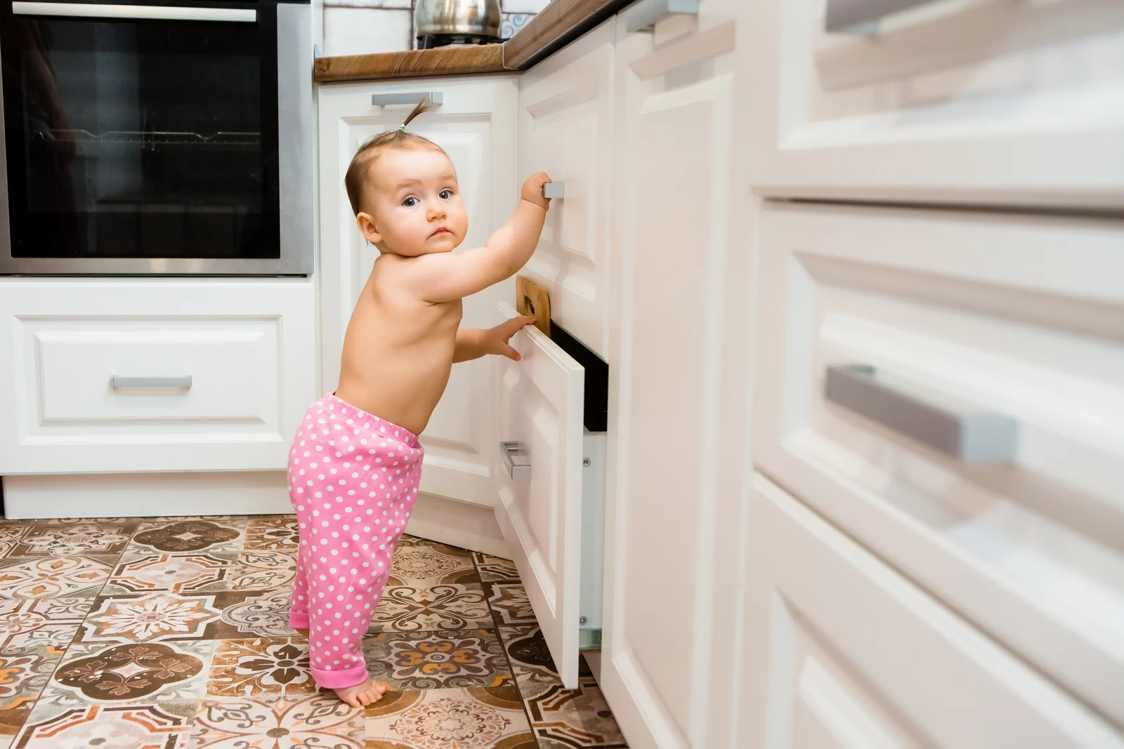 A baby in pink polka-dot pants stands on a tiled kitchen floor and pulls open a low white cabinet door