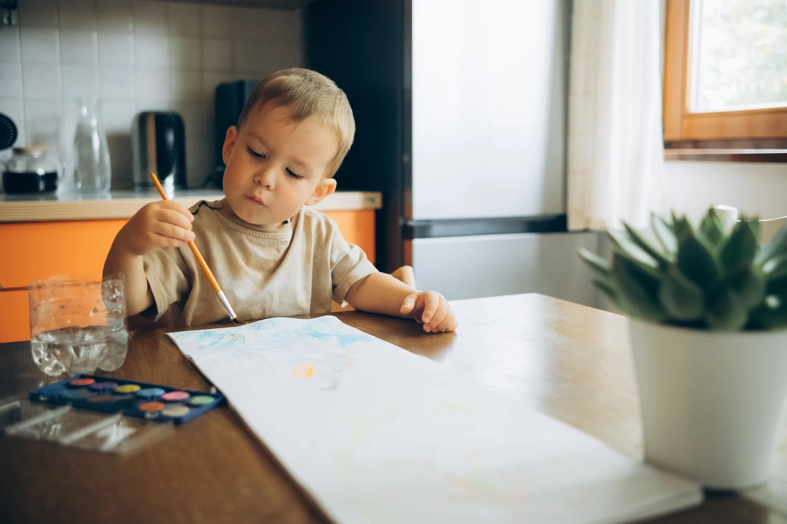 Toddler painting with a brush on large paper at a wooden kitchen table, with a watercolor set and a glass of water