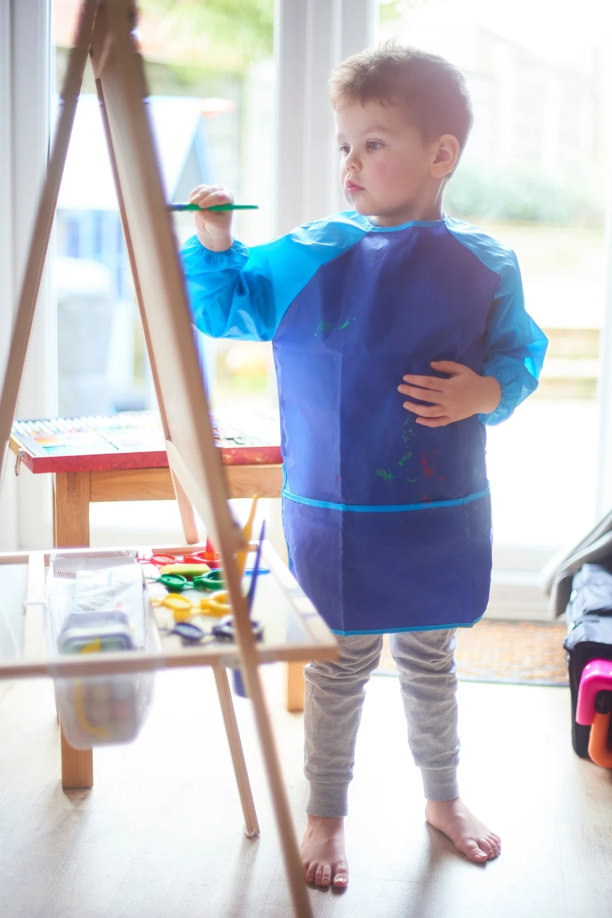 Toddler in a blue smock painting at a small easel by a sunny window, paint pots on a low stool beside it