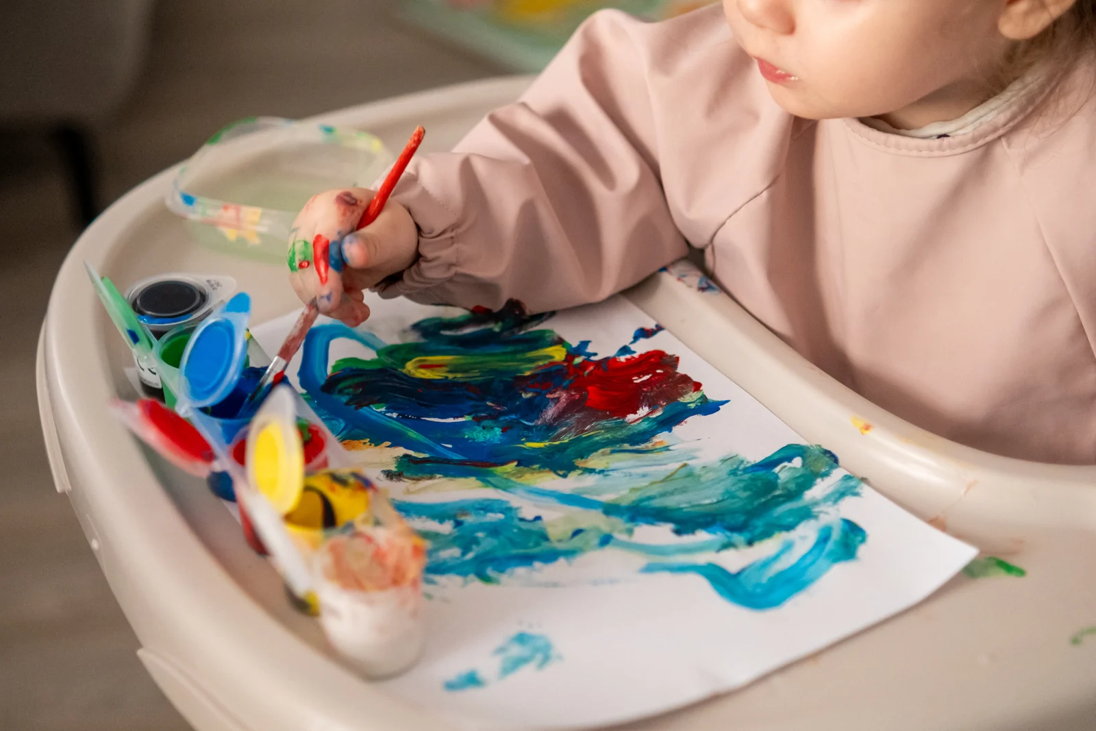 Toddler in a high chair painting on paper with a brush, colors swirled across the page