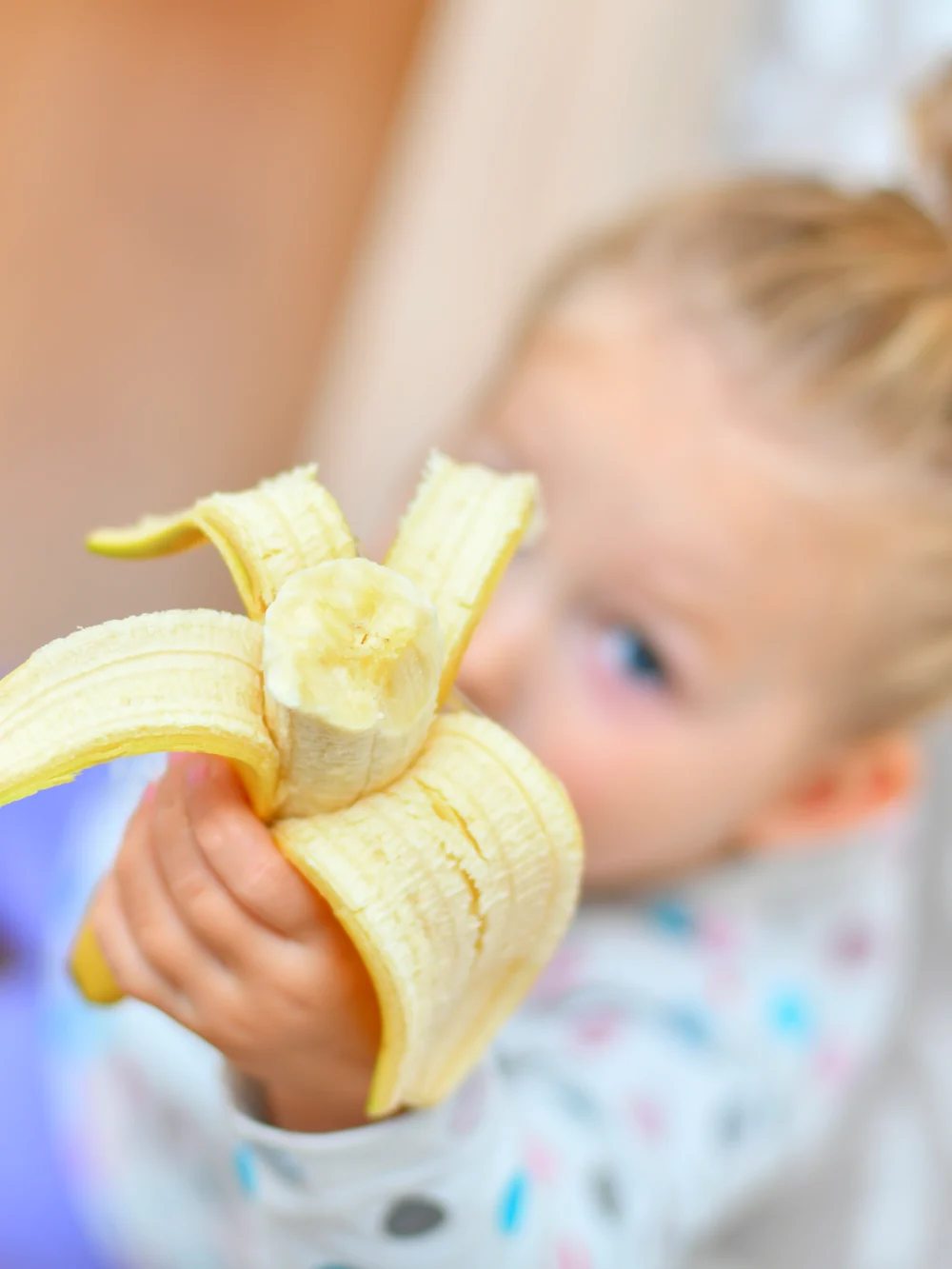 Toddler with a hair bun holding up a half-peeled banana, face softly out of focus behind it