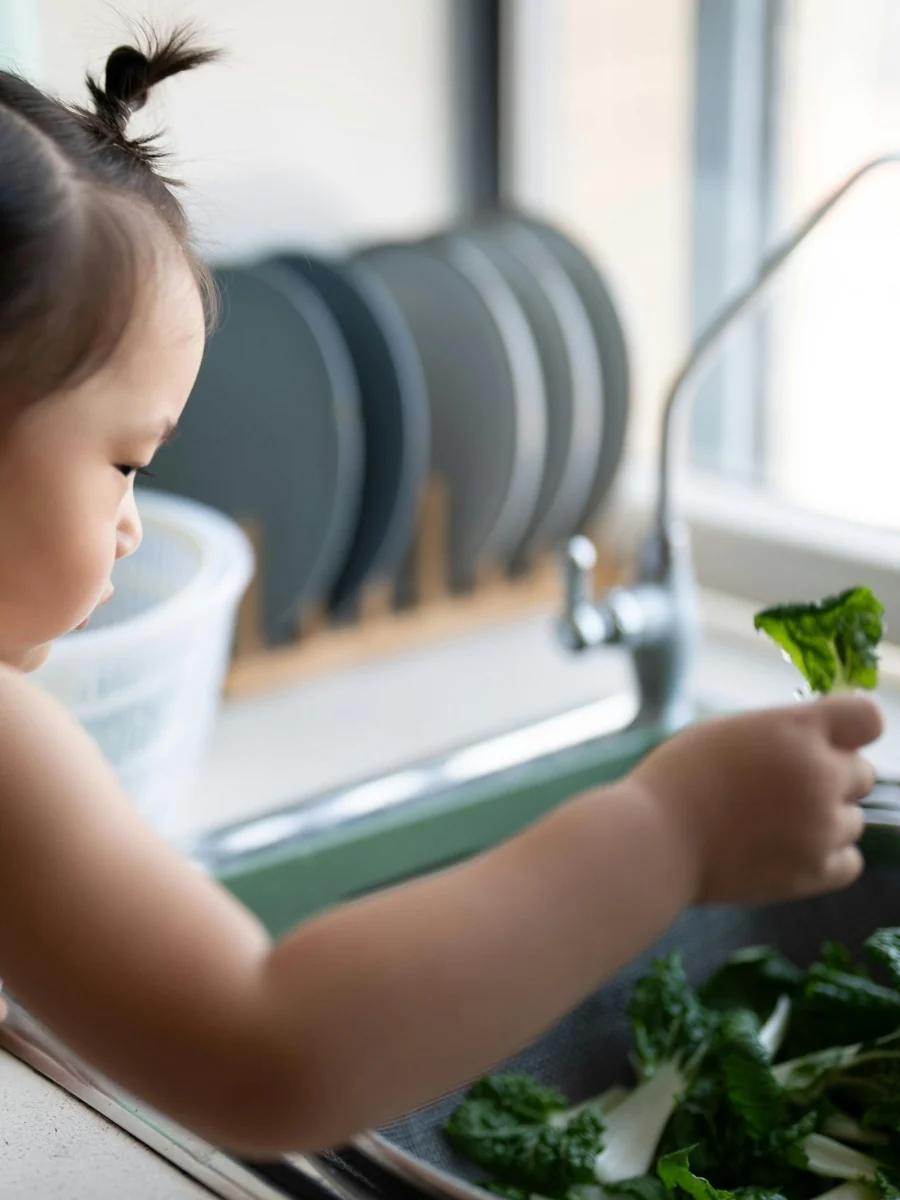 Toddler at the kitchen sink holding up one leaf from a pile of leafy greens