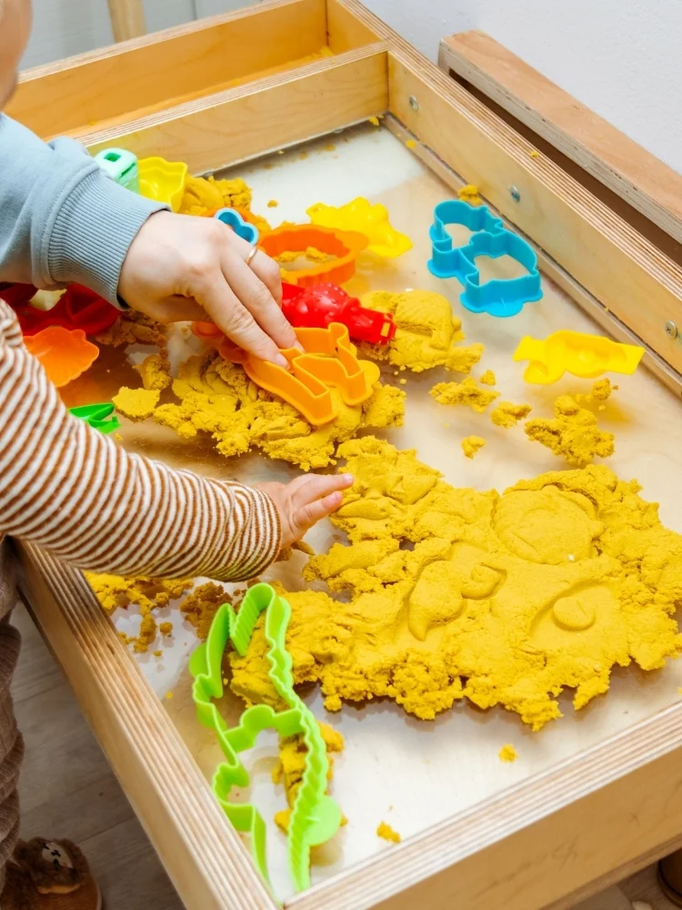 Toddler pressing yellow dough in a wooden tray with raised edges, plastic shapes beside it