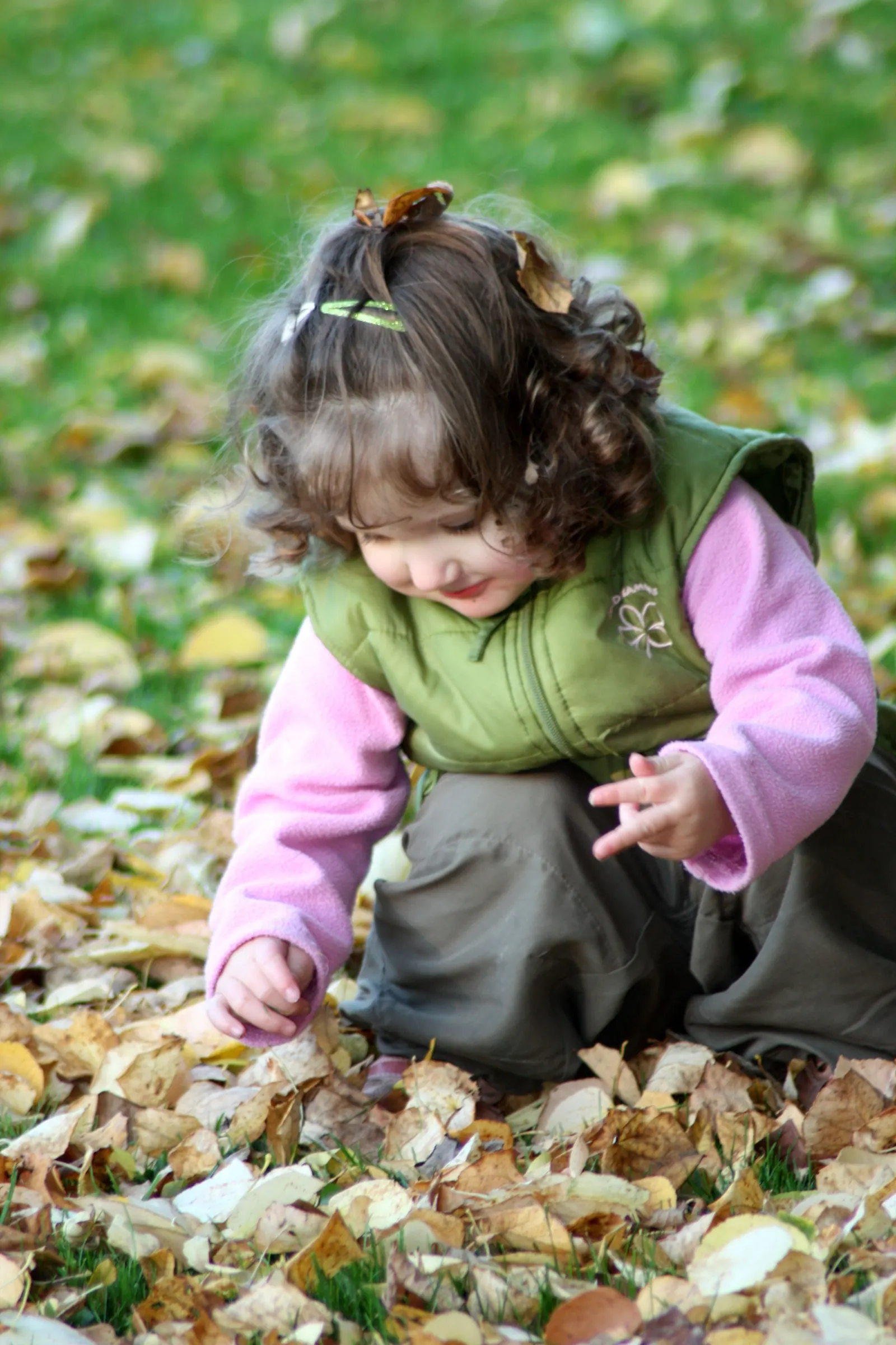 A toddler in a green vest crouches in a pile of fallen leaves outdoors, sorting through them