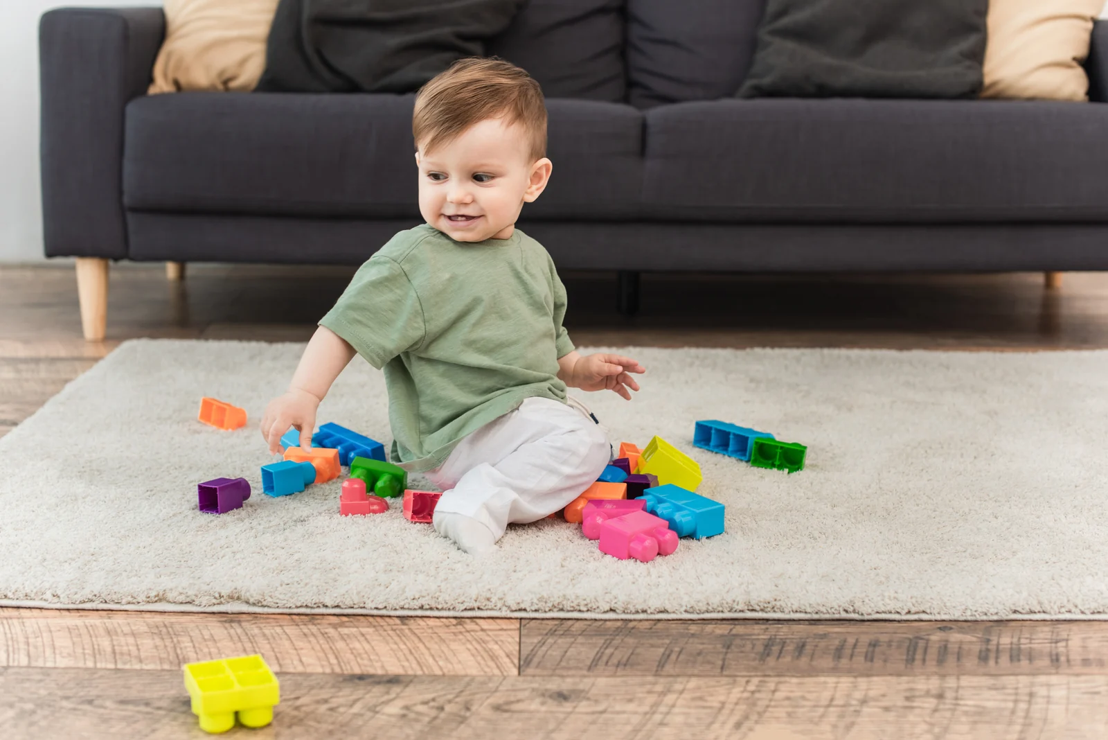A toddler sitting on a cream rug in front of a gray sofa, playing with chunky colorful plastic building blocks