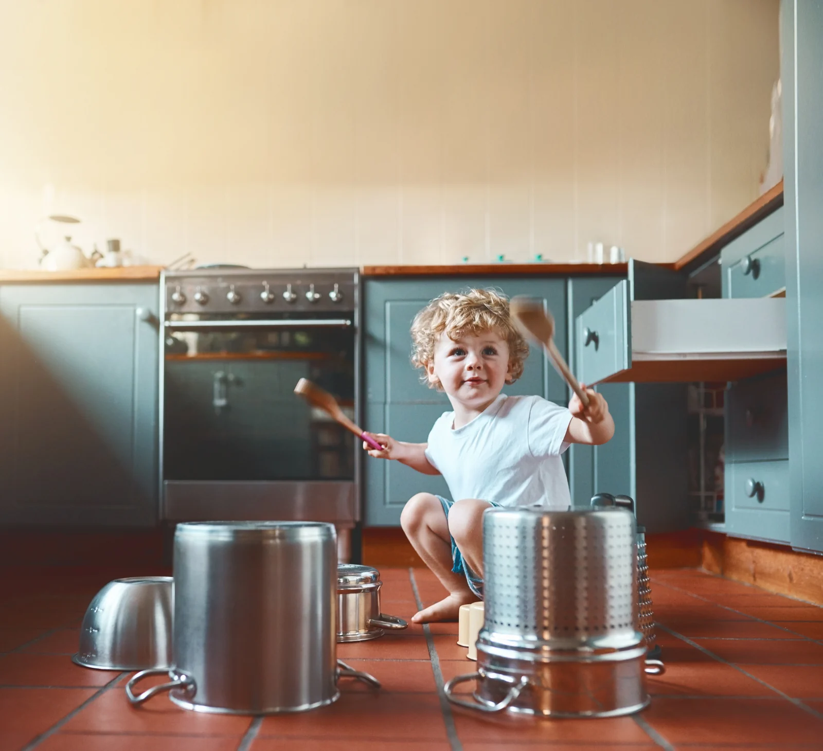Toddler crouching on a kitchen floor, drumming on steel pots and a bowl with two wooden spoons